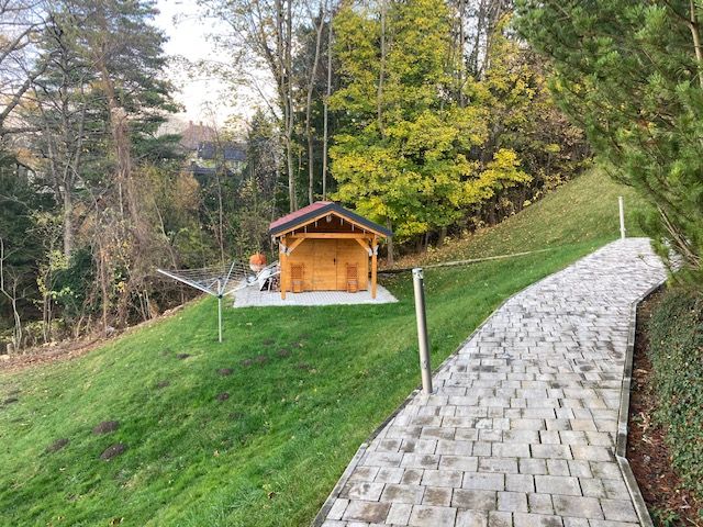 A garden with a small wooden house, surrounded by trees and a paved path.