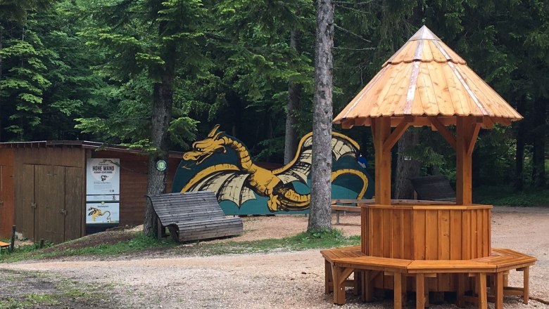A wooden pavilion in a forest square with a painted dragon in the background.