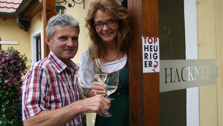 A man and a woman are standing in front of a building with a sign saying 'Top Heuriger' and holding wine glasses.