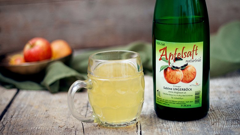 A bottle of apple juice and a glass filled with apple juice on a wooden table, with a green cloth in the background.
