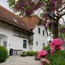 A country house with red roof tiles, surrounded by blossoming trees and pink flowers in the foreground.