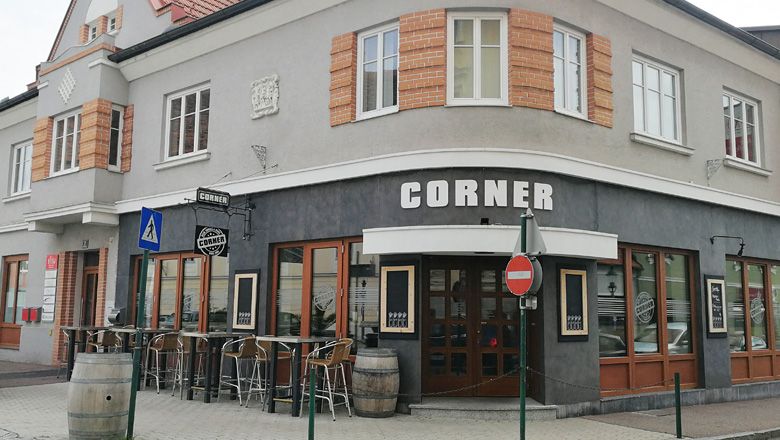 Corner bar with gray facade and wooden doors, tables and barrels outside.