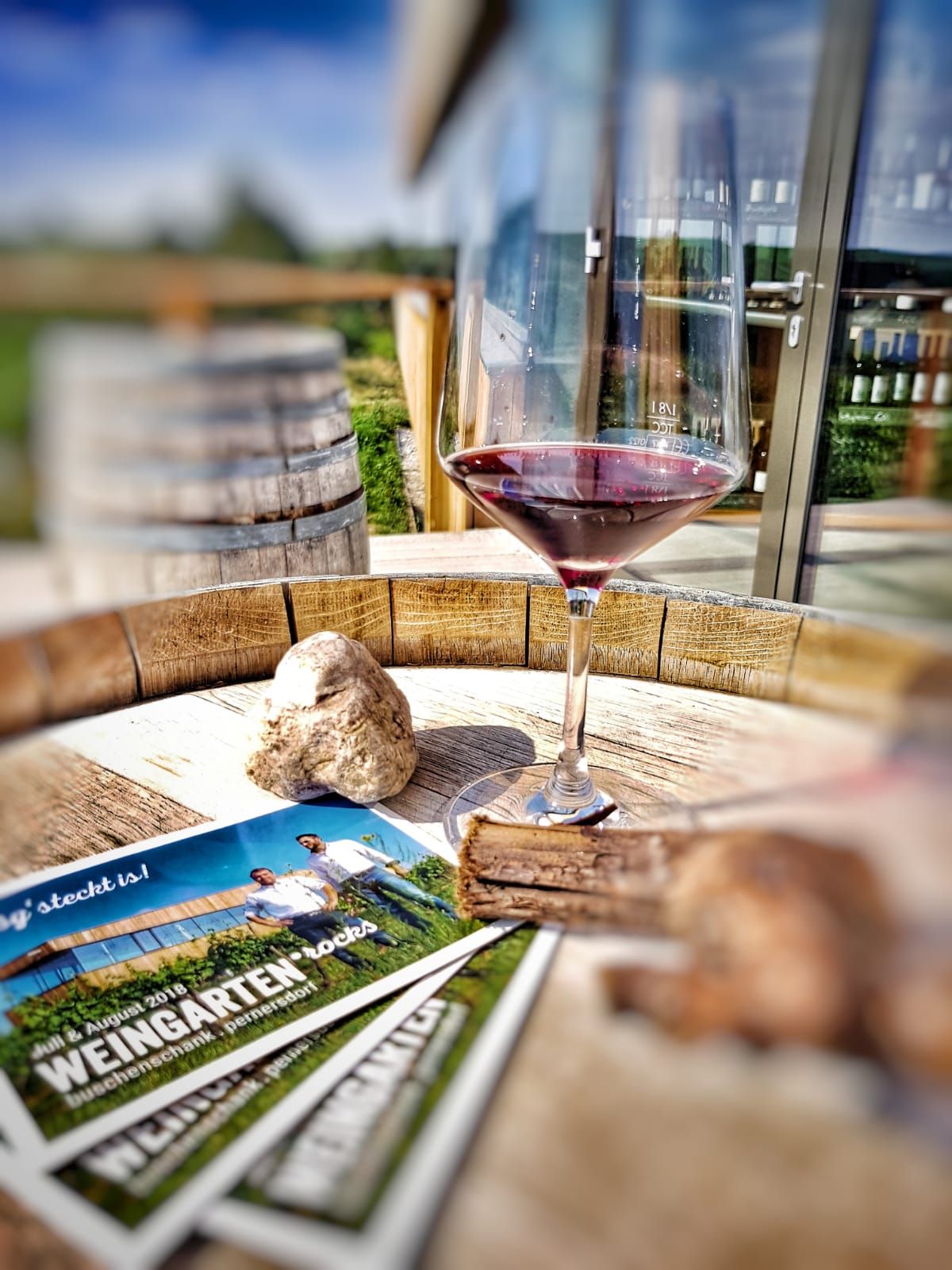A wine glass with red wine stands on a wooden table next to brochures and a stone.