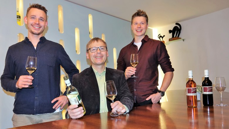 Three men are standing at a table with wine glasses and bottles of wine.
