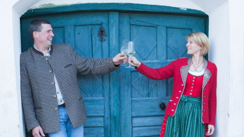 Schönhofer family, © Sabine Schneider A man and a woman clink glasses of wine in front of a blue wooden door.