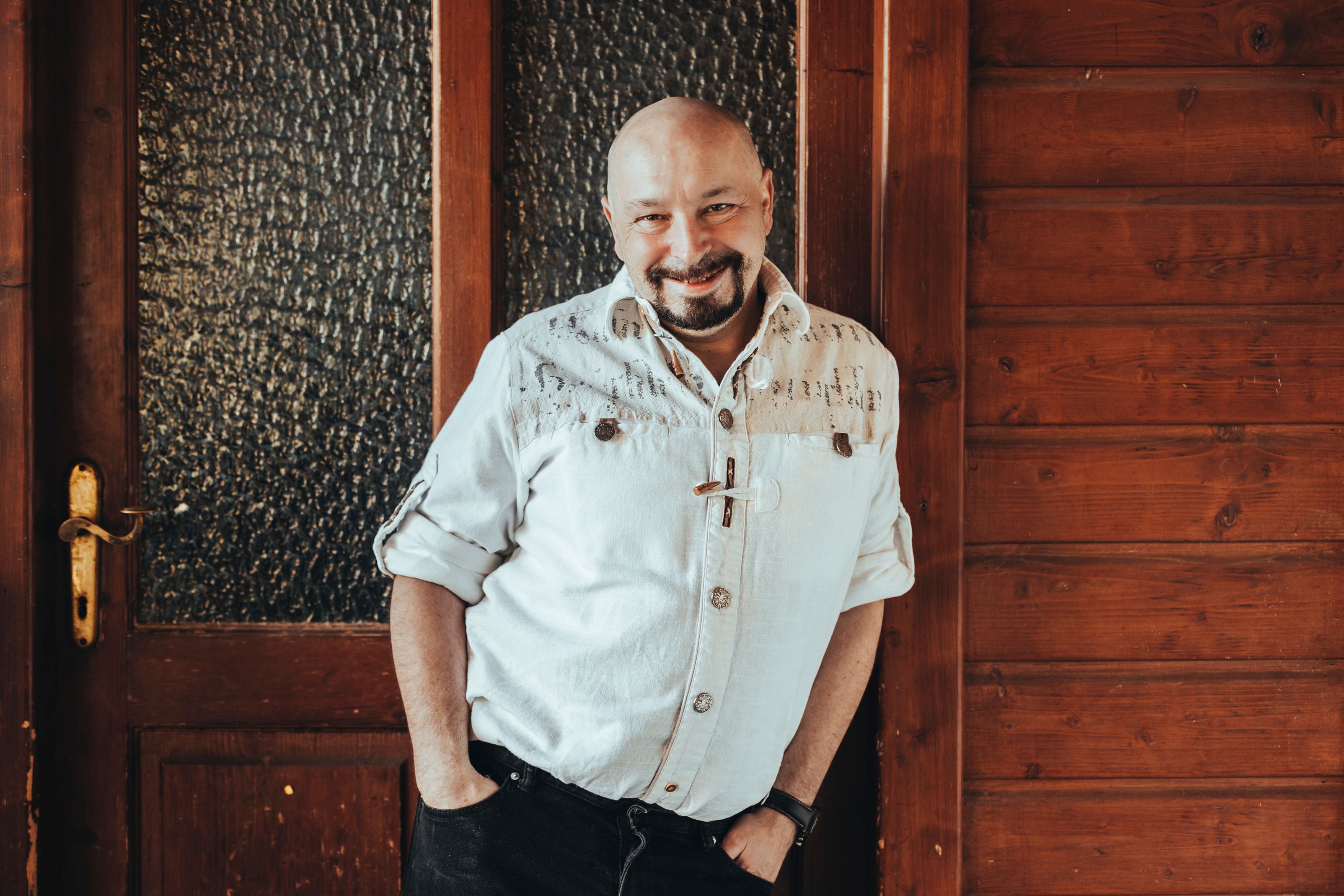 A man in a white shirt leans against a wooden wall, smiling.