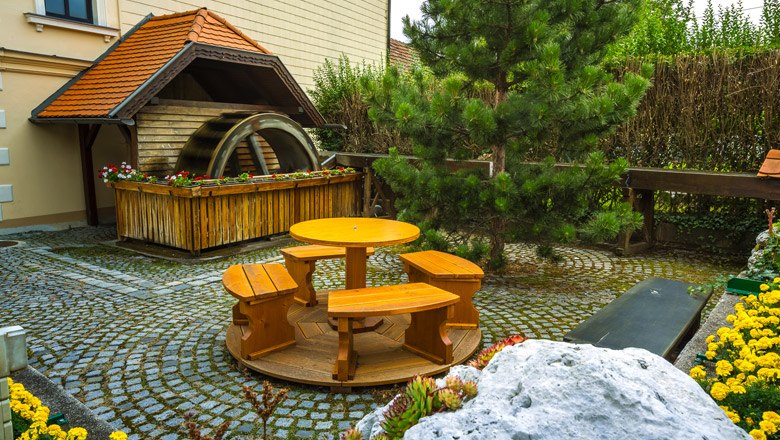 A small water wheel next to a wooden table with benches in a paved garden.