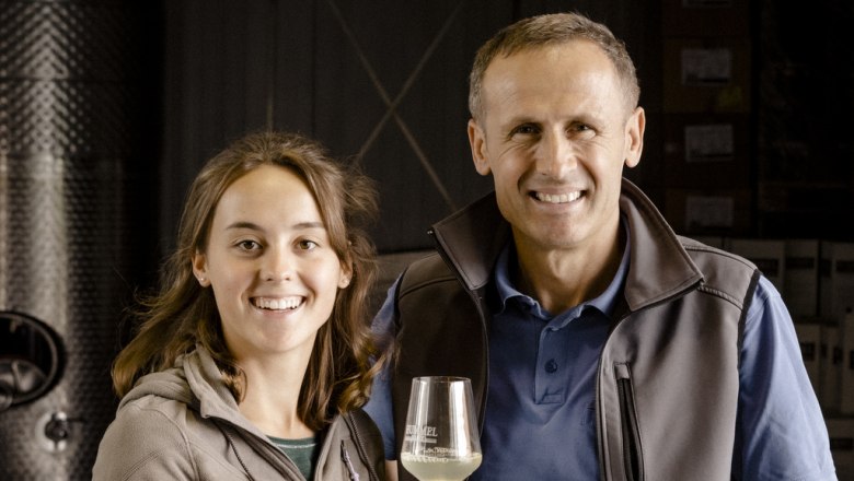 A man and a woman smile at the camera, the man holds a wine glass. They are standing in a wine cellar.