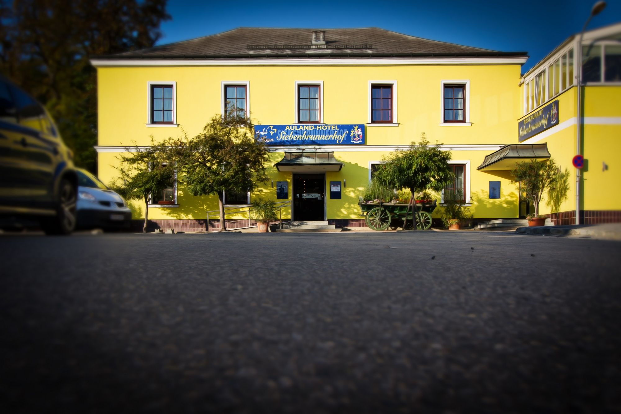 Yellow building with the inscription 'Auland-Hotel Siebenbrunnerhof', surrounded by trees and an old car.