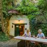 Two people are sitting at a wooden table in front of a wine cellar in the countryside.