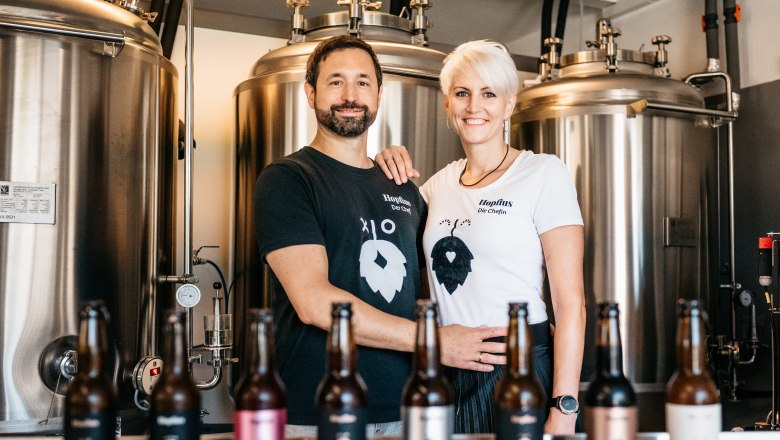 A couple stands in front of brewery tanks with beer bottles in the foreground.