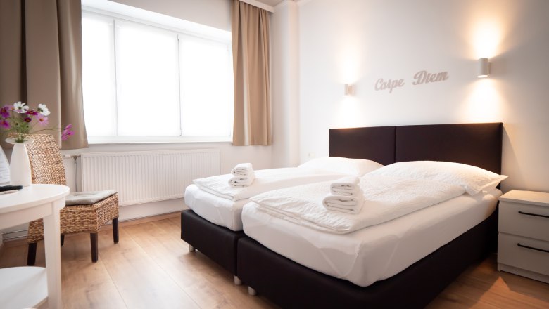 Double room, © Doris Eminger Modern double room with two beds, white comforters and towels. 'Carpe Diem' lettering on the wall, light-colored curtains and wooden floor.
