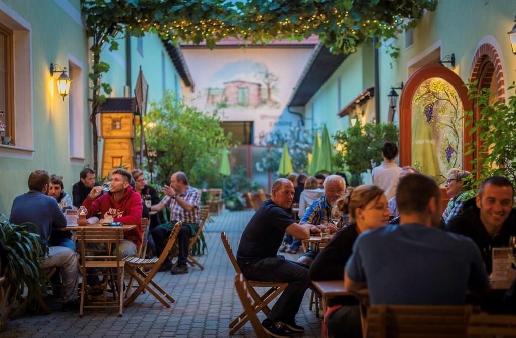 People sit at wooden tables in a cozy courtyard of a winery, surrounded by plants and fairy lights.