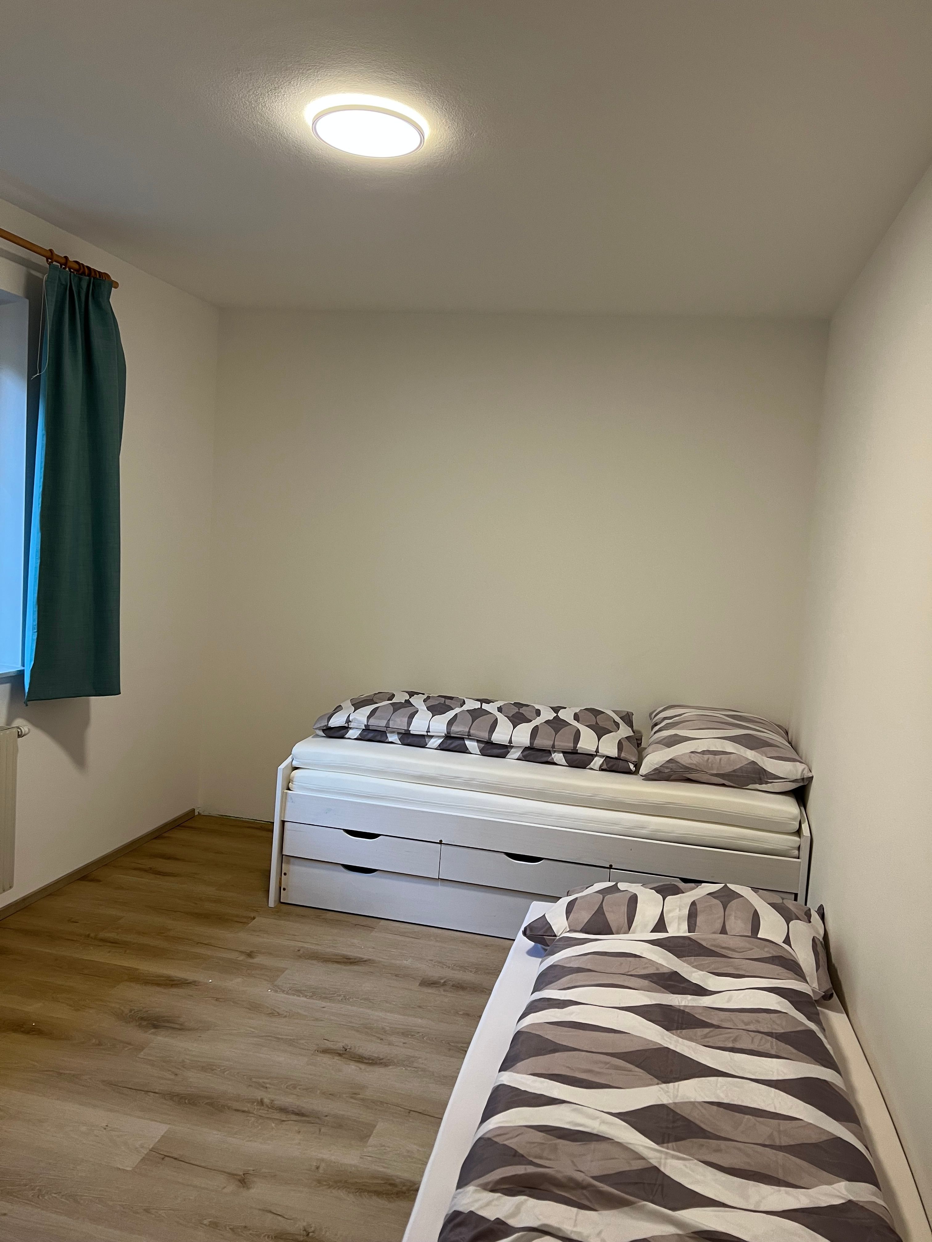 A simply furnished twin room with a wooden floor, two single beds and a window with blue curtains.