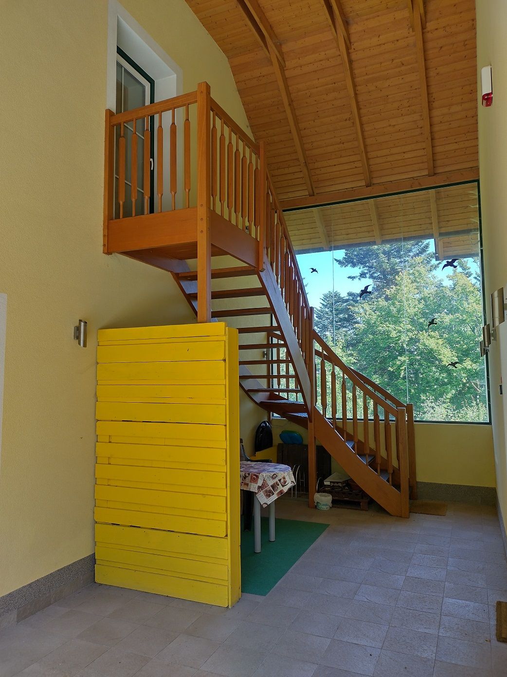 Wooden staircase in an interior with a yellow wall and large window with a view of trees.