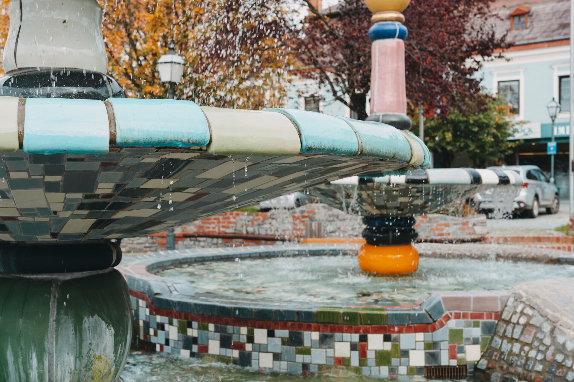Der Herbst verleiht dem Hundertwasserbrunnen eine besondere Magie, während das Wasser sanft über die bunten Fliesen plätschert. Umgeben von leuchtenden Bäumen und einer einladenden Atmosphäre, lädt dieser Ort zum Verweilen und Träumen ein.