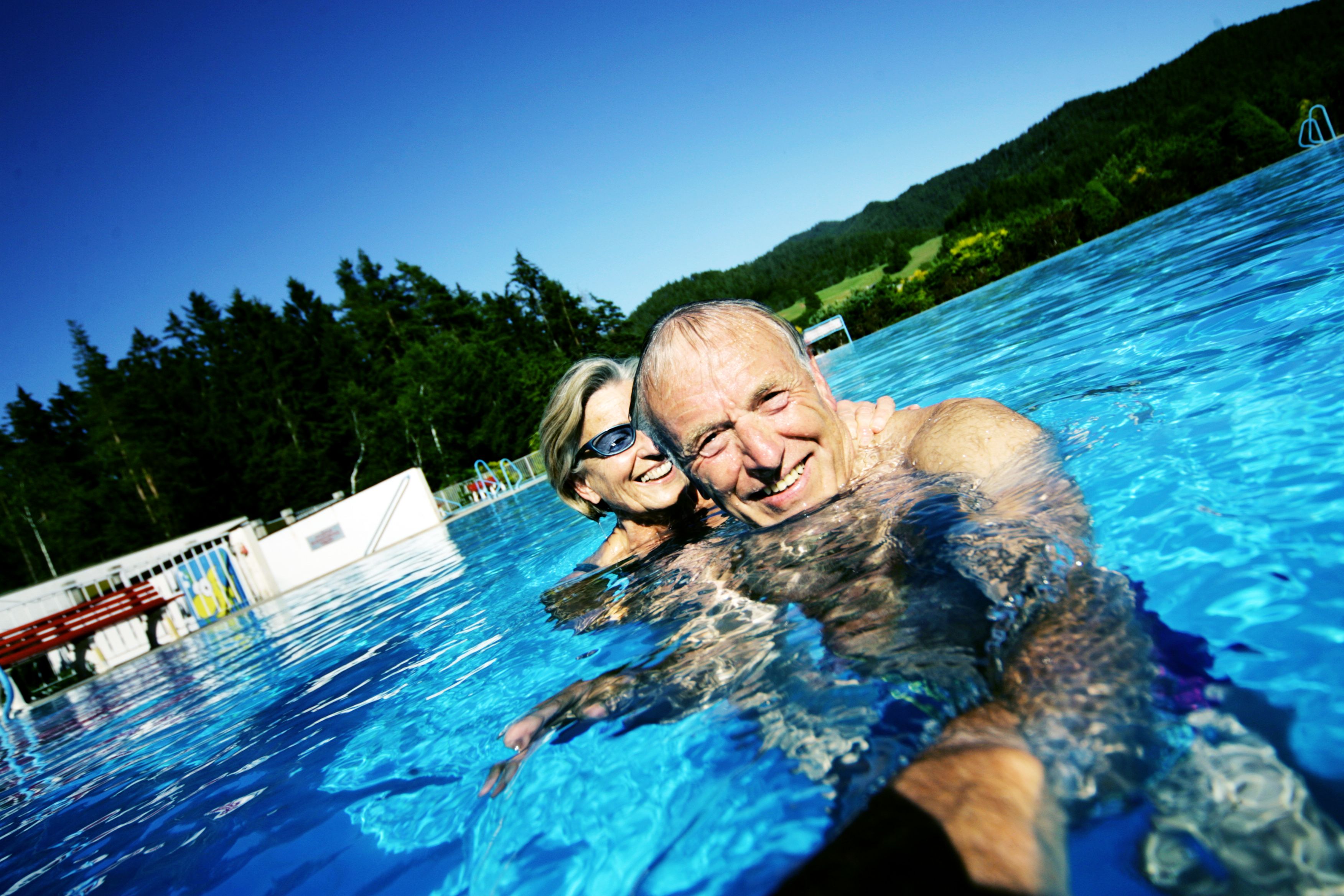 An elderly couple smiling as they swim in an outdoor pool with a forest in the background.