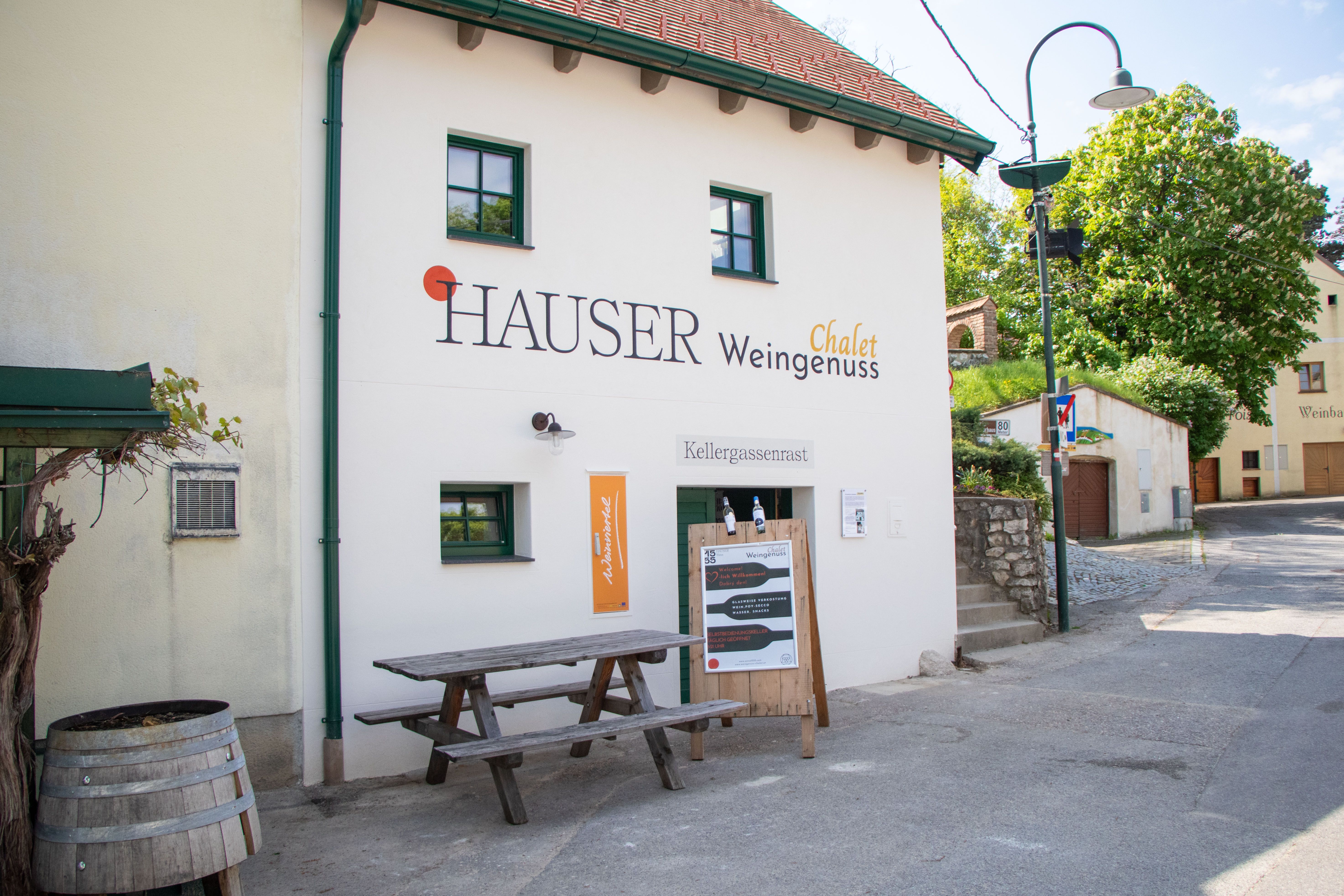 Exterior view of a building with the inscription 'Hauser Chalet Weingenuss'. In front of the building is a wooden table and a wine barrel.