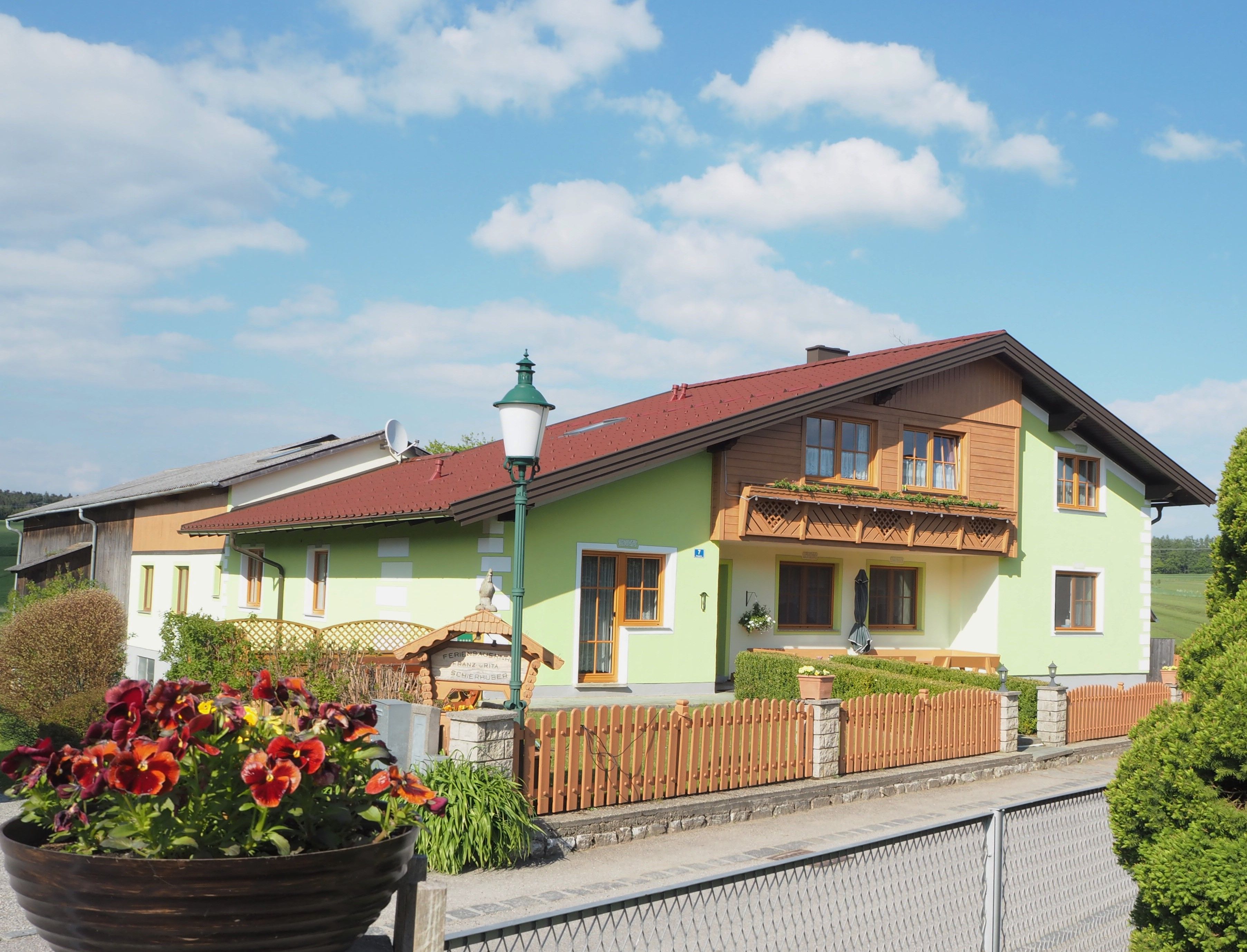 A vacation farm with green paint and wooden balcony, surrounded by a wooden fence and flowers in the foreground.