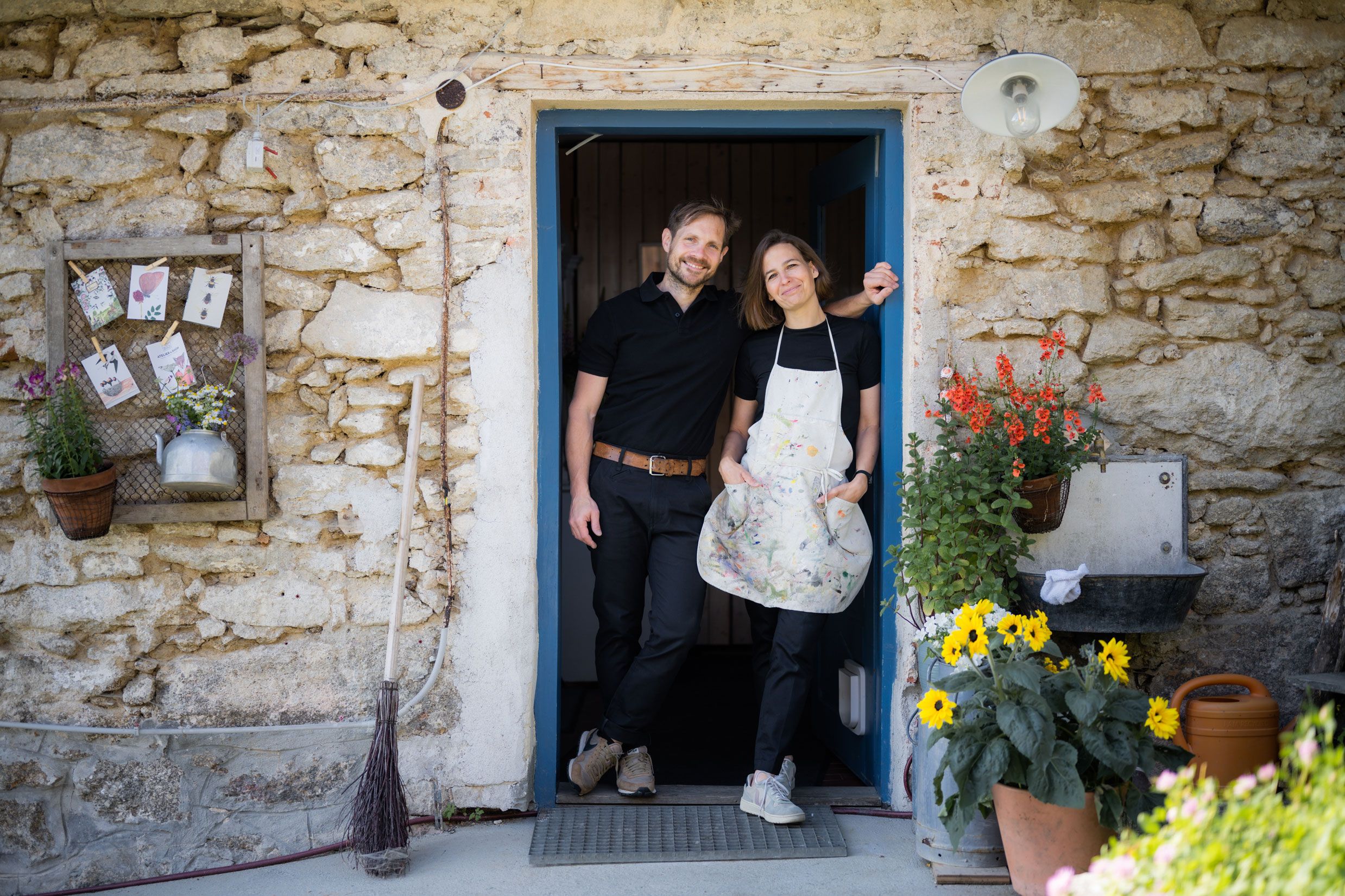 A man and a woman stand smiling in a doorway of a rustic stone house, surrounded by flowers and decorations.
