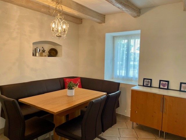 A cozy dining area with a wooden table, black chairs and a corner bench. A chandelier hangs on the wall, next to it a window and a sideboard.