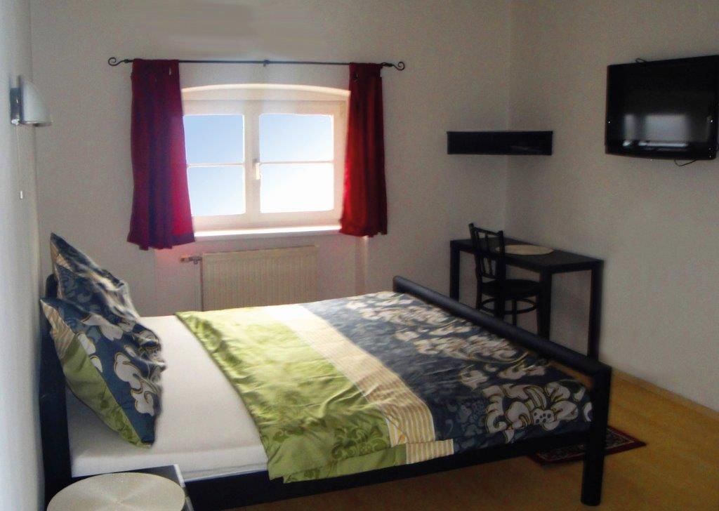 A bedroom with a bed, a window with red curtains, a small table and chair and a TV mounted on the wall.
