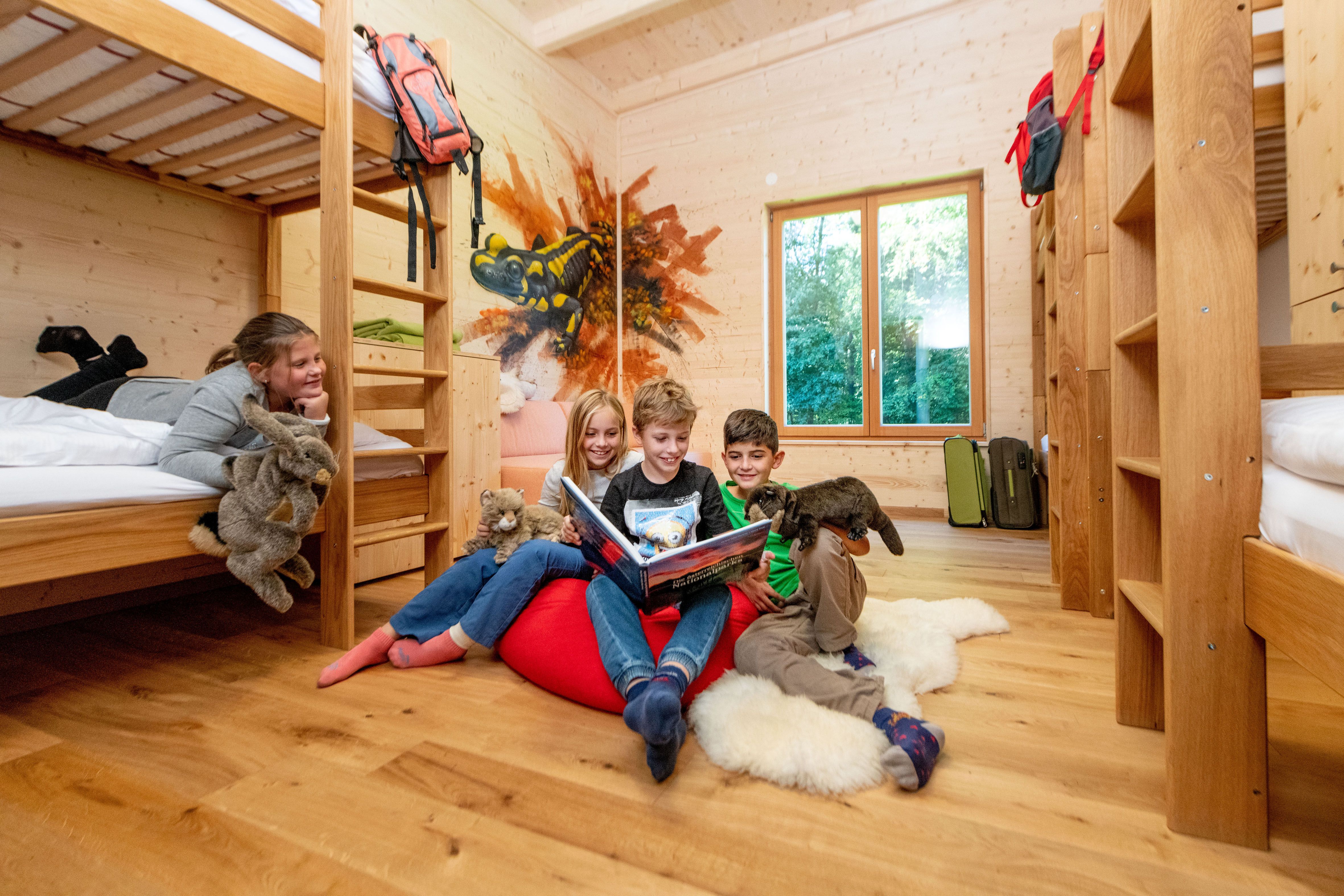 Children in a wooden room with bunk beds, reading a book and holding cuddly toys.