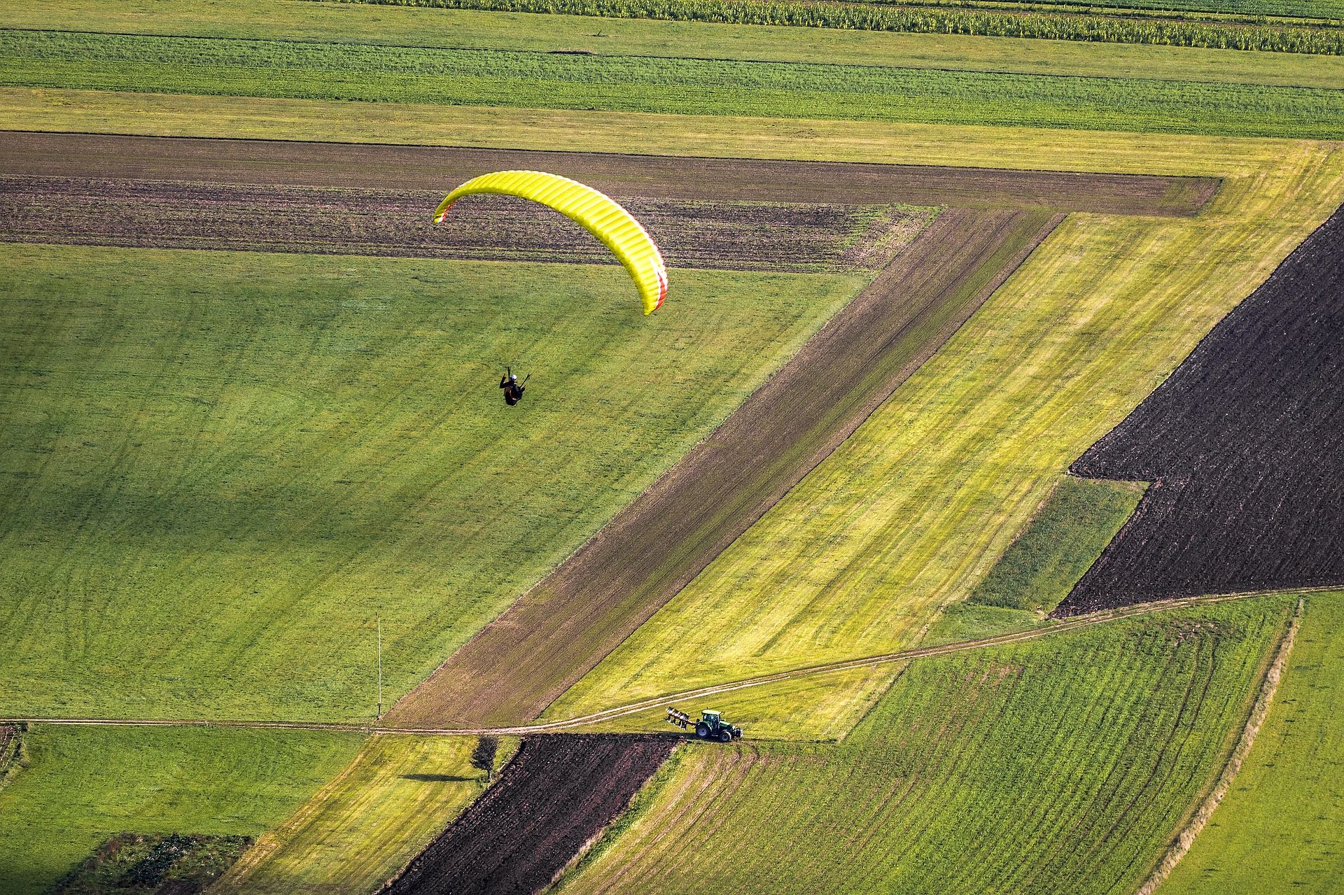 A parachutist with a yellow parachute over green fields, below a tractor on a dirt road.