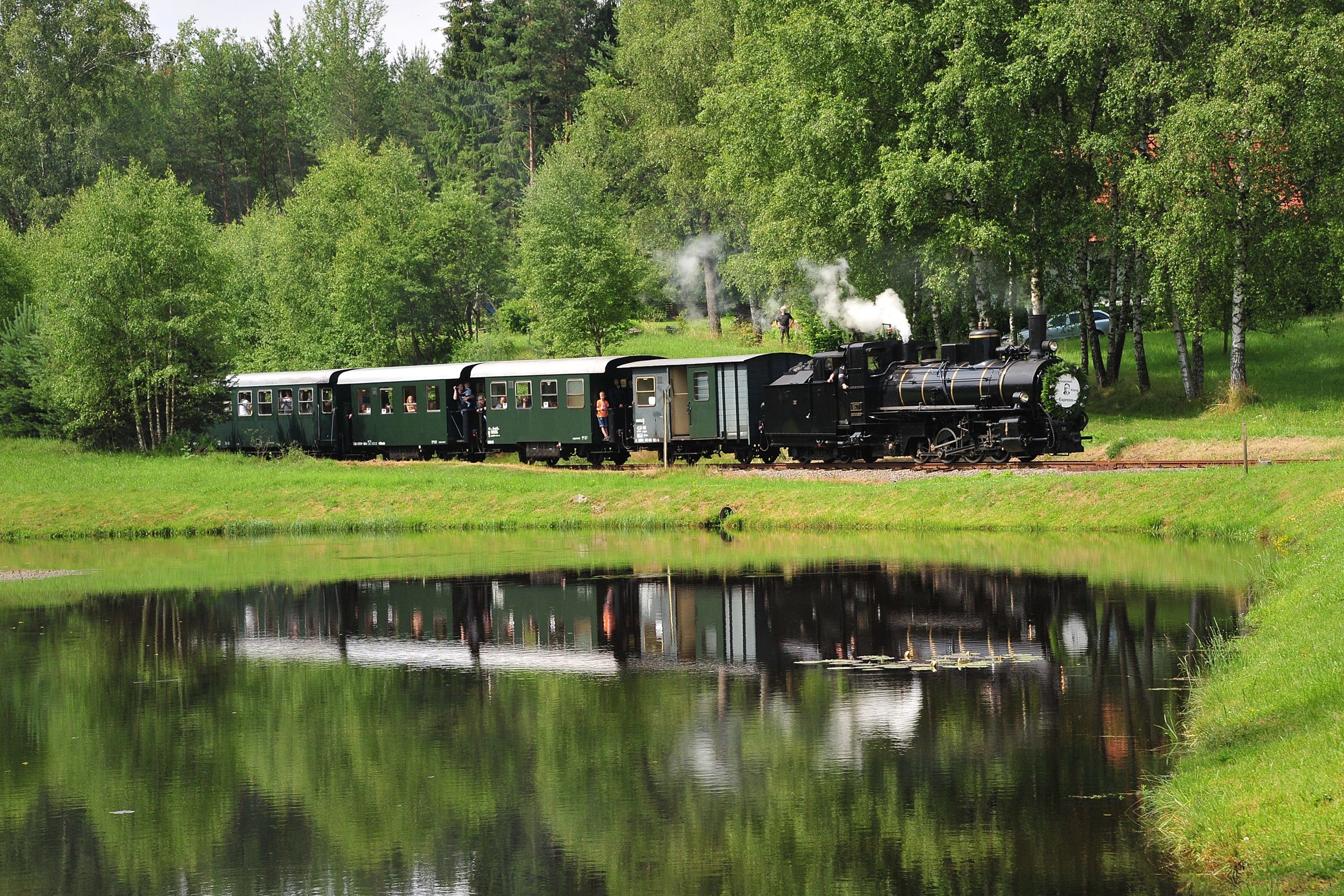 A historic steam locomotive pulls green wagons through a verdant landscape reflected in a tranquil pond.