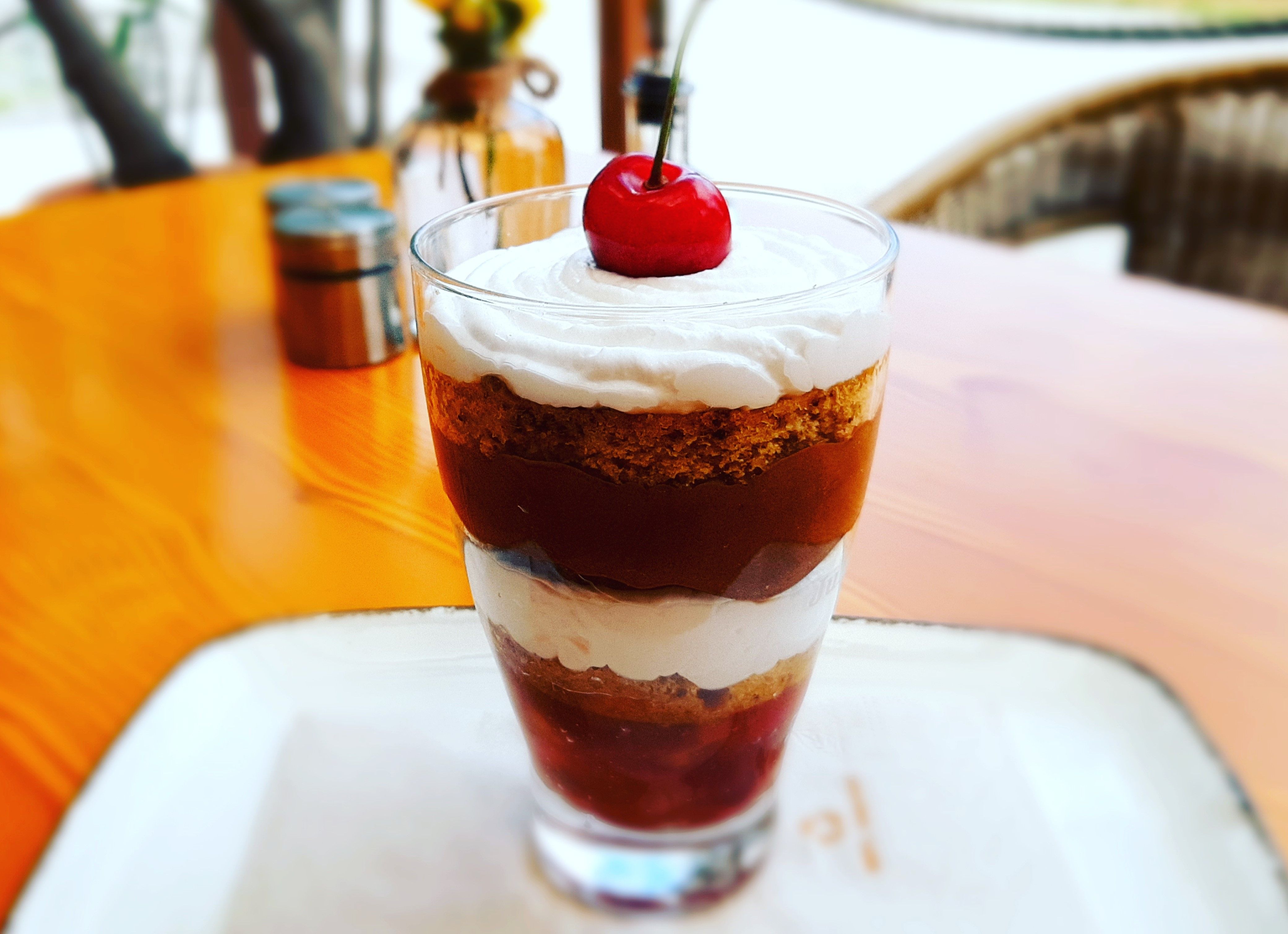 A dessert in a glass with layers of cream, cake and cherries, garnished with a cherry.