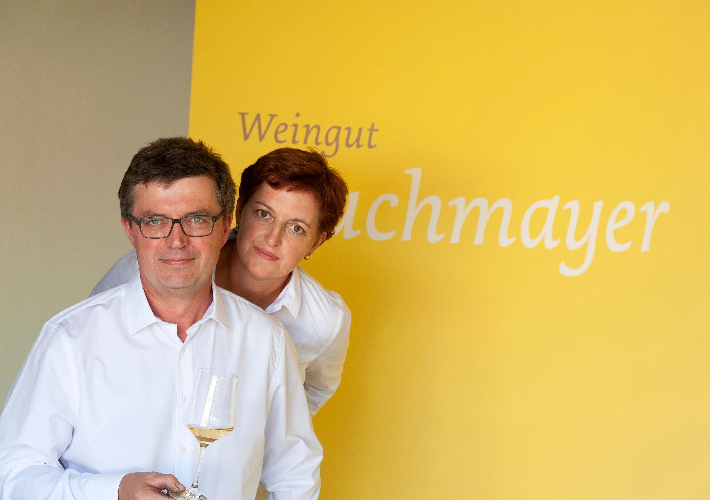 Two people in white shirts in front of a yellow background with the inscription 'Weingut Buchmayer'. One person is holding a wine glass.