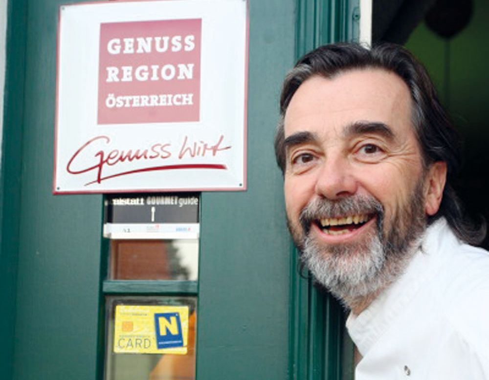 A man with a beard smiles in front of a sign reading 'Genuss Region Österreich'.