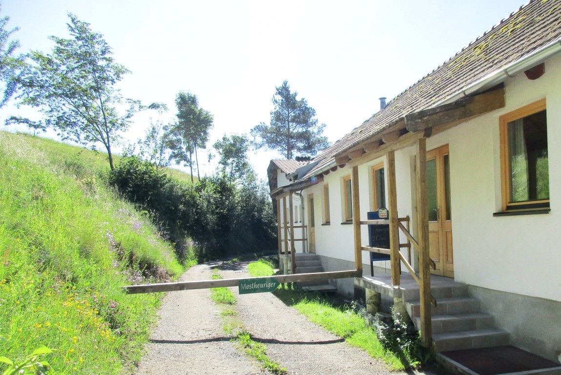 White building with a barrier with the sign "Tavern" next to a gravel path and a green meadow with trees.