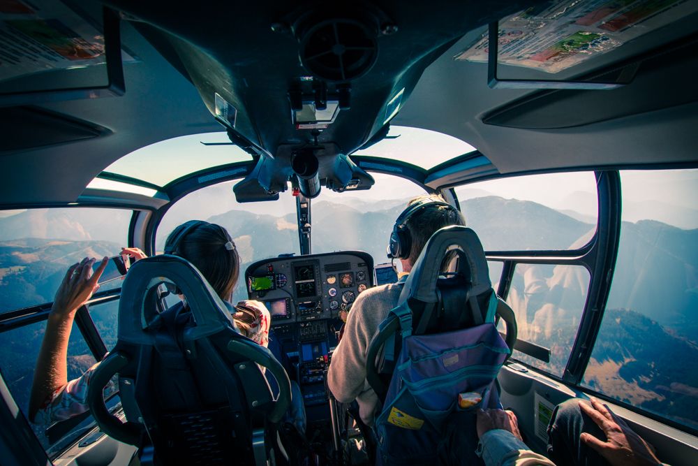 View from the cockpit of a helicopter with two people flying over a mountain landscape.