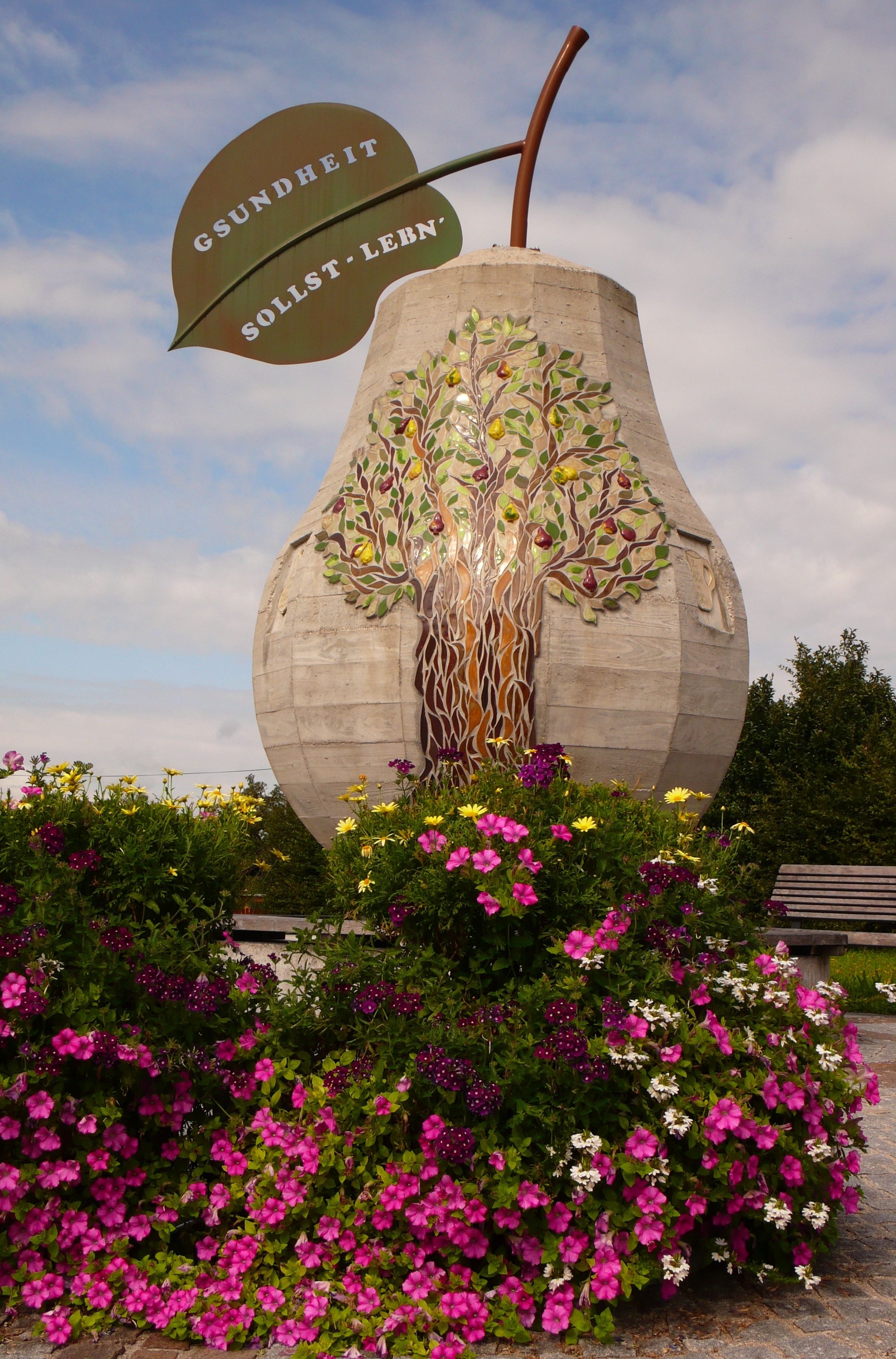 Large concrete bulb with leaf inscription 'Gsundheit sollst lebn' and flowers in the foreground.