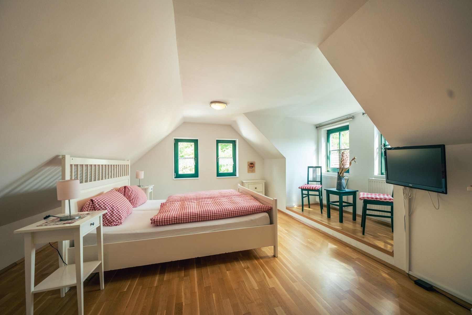 Bright bedroom with sloping ceiling, double bed, wooden floor, two windows, table with chairs and TV on the wall.