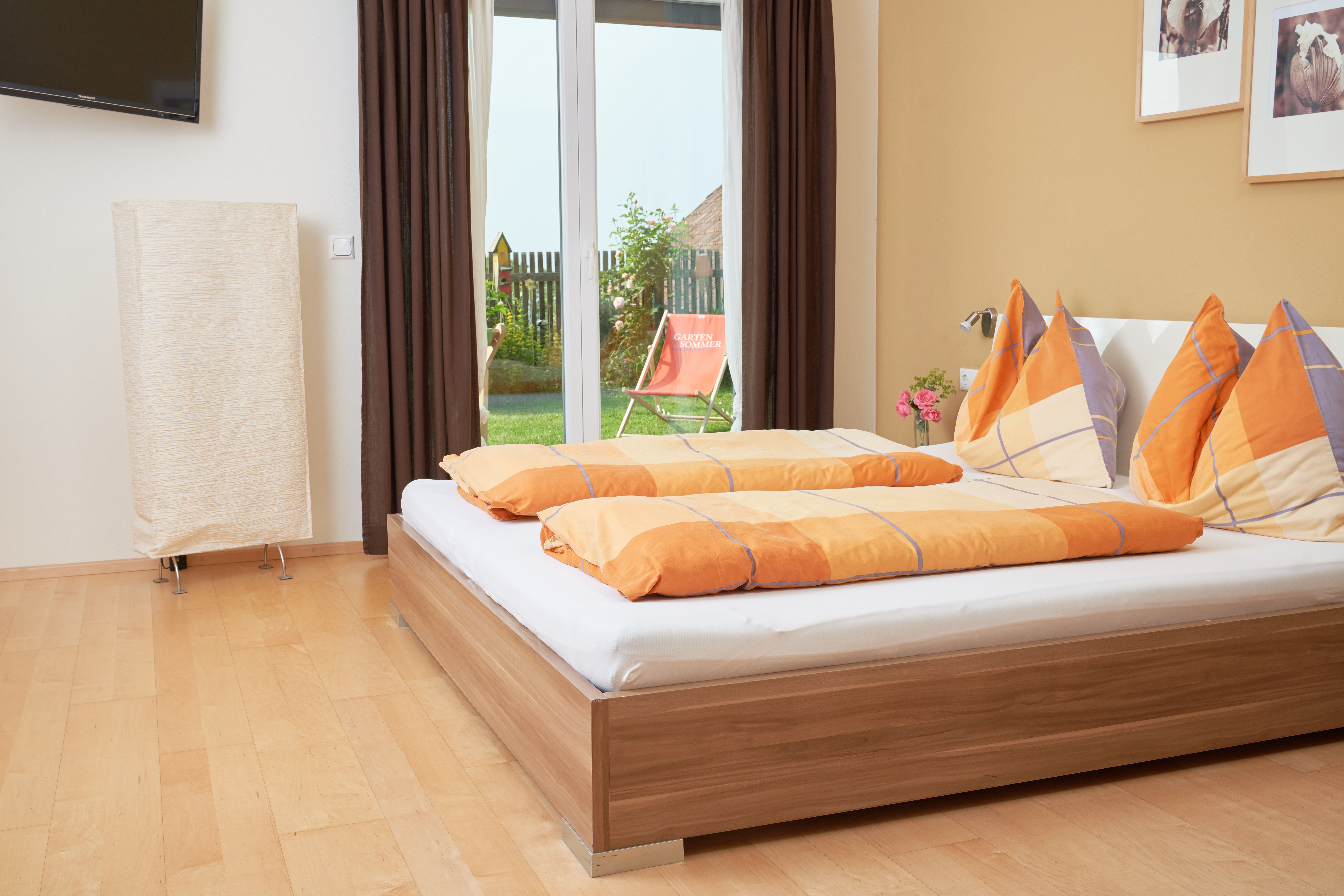 A cozy bedroom with a double bed, orange comforters and a view of a garden through a glass door.