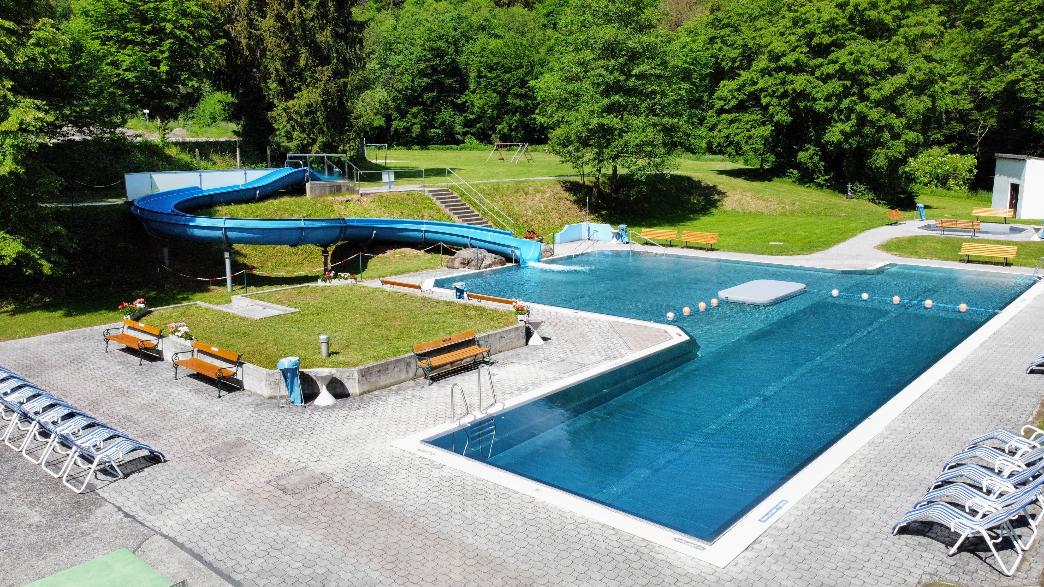 An outdoor pool with slide and sun loungers, surrounded by trees.
