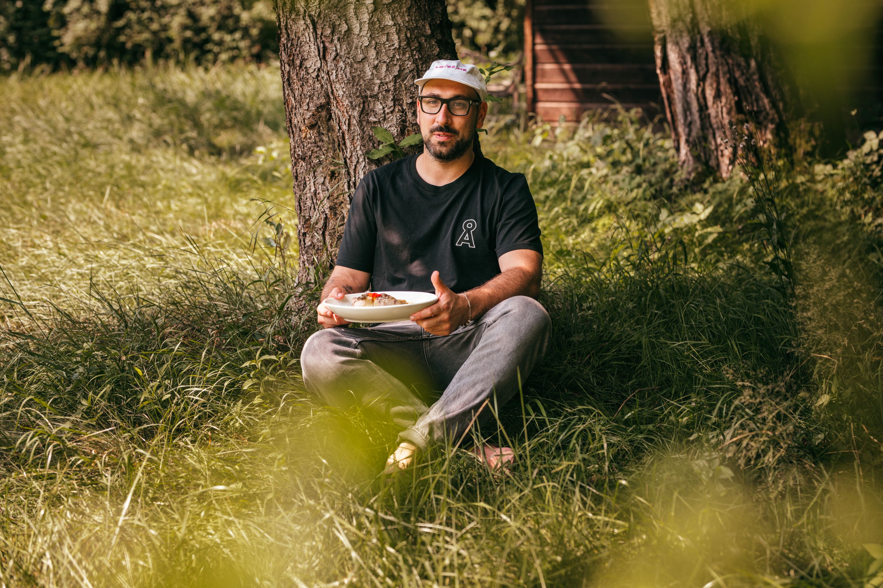 Man sitting in the grass under a tree holding a plate of food.