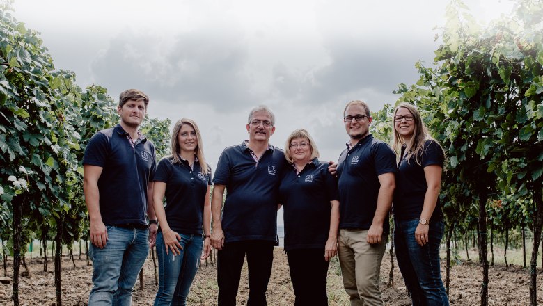 A family is standing in a vineyard, all wearing dark blue polo shirts with a logo.