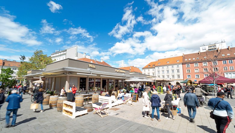 A lively market square with people lingering at stalls and cafés under a blue sky.