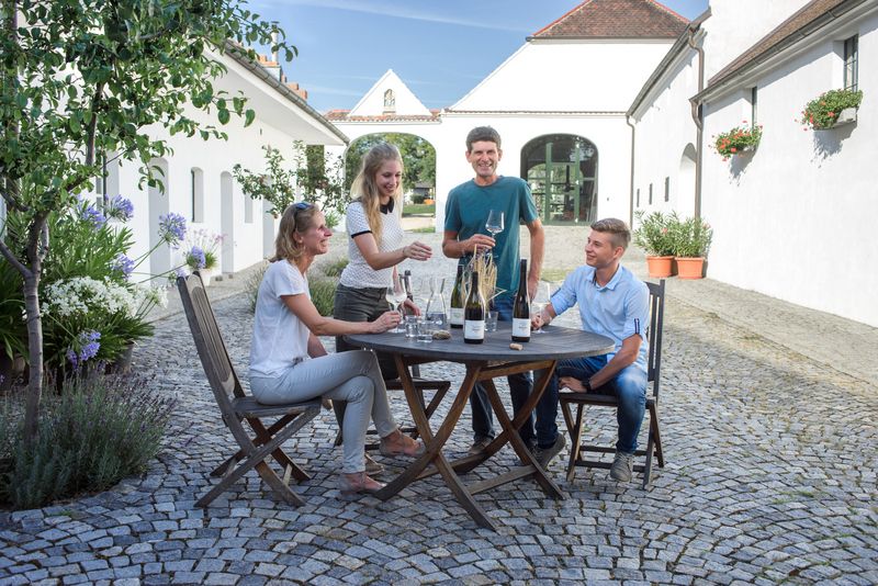 Four people sit at a round table outside and enjoy wine. They are surrounded by white buildings.