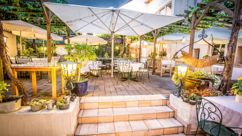Cozy guest garden with tables, chairs and parasols, surrounded by plants and lights.