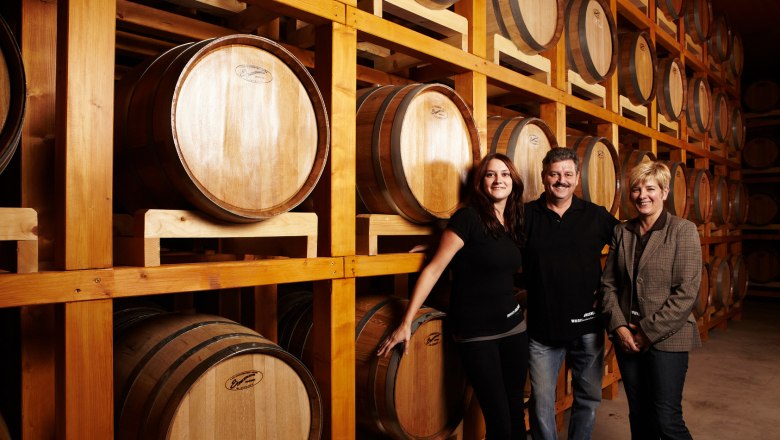 Three people stand in front of wooden barrels in a wine cellar.