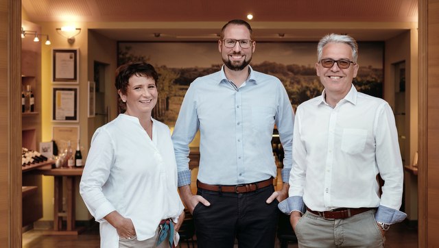 Three people in a room with bottles of wine in the background.