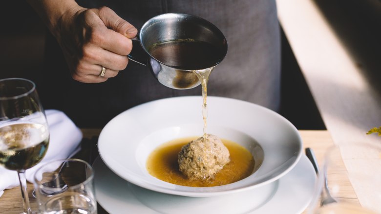 A person pours broth over a liver dumpling in a white plate.