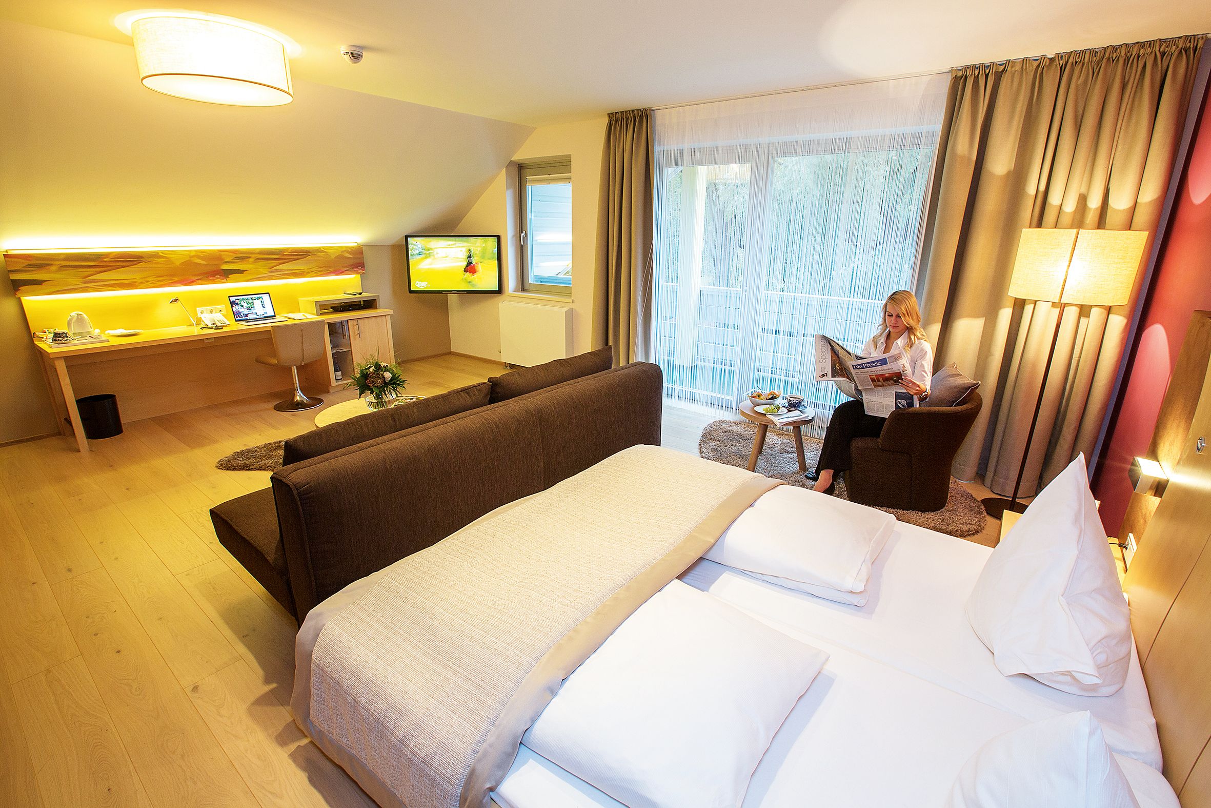 Modern hotel room with double bed, desk, TV and a woman reading the newspaper.