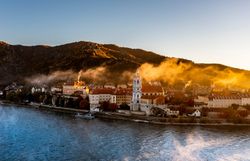 Die sanften Hügel der Wachau umarmen das malerische Stift Dürnstein, während die goldenen Strahlen der Herbstsonne die Landschaft in ein warmes Licht tauchen. Nebelschwaden ziehen über die Donau und verleihen der Szenerie eine mystische Atmosphäre, die zum Verweilen einlädt.