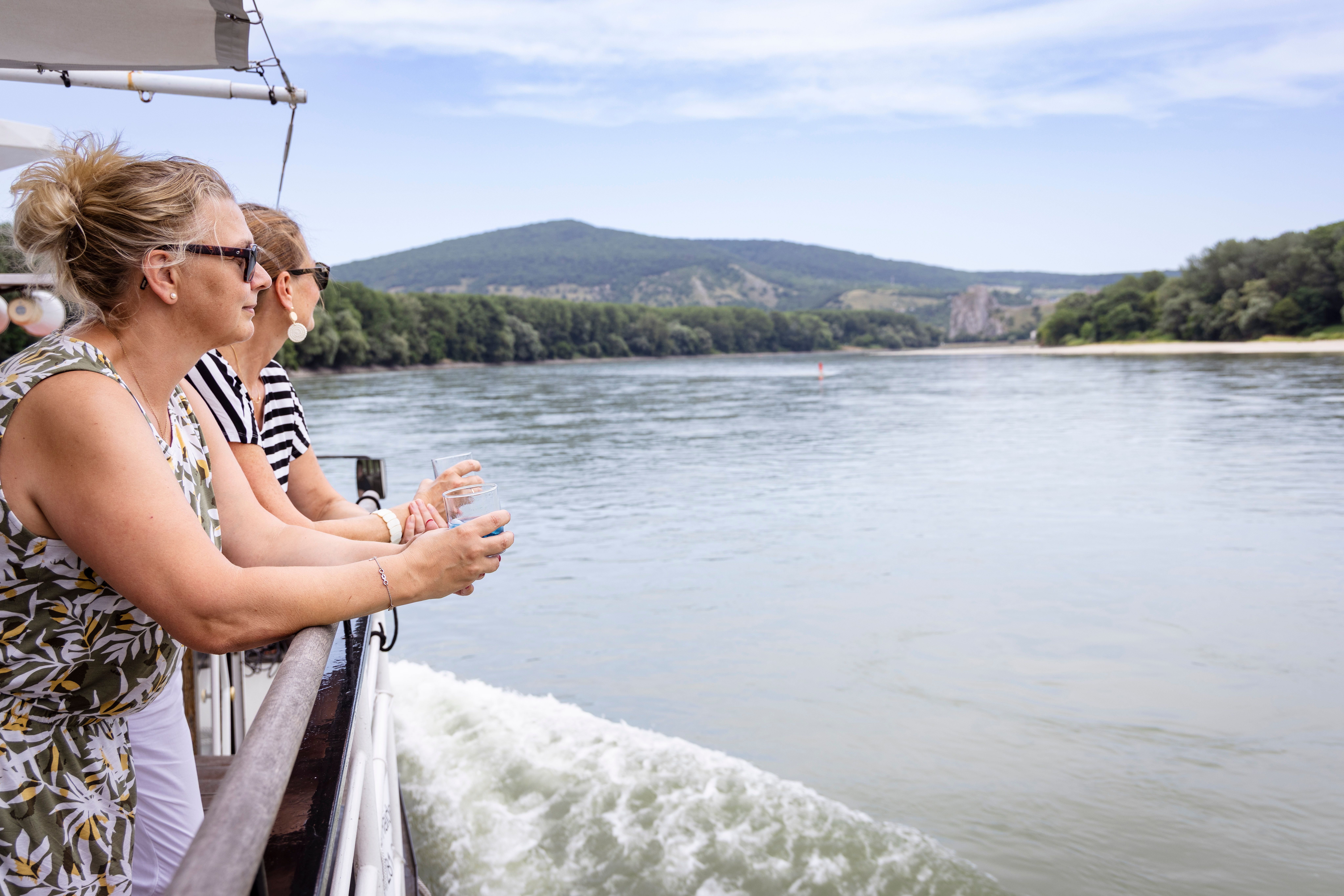 Two women stand at the railing of a boat on the Danube and enjoy the view of the surrounding landscape.