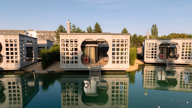 Therme Laa | Hotel | Silent Spa | Villas, © Niederösterreich Werbung / Maximilian Pawlikowsky Modern villas on water with reflective facades and stairs, surrounded by nature.